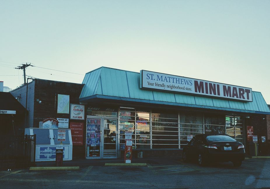 A storefront of St. Matthews Mini Mart, a local convenience shop with a blue metal-sloped roof and large glass windows displaying various products. In front of the store, a black car is parked on the 