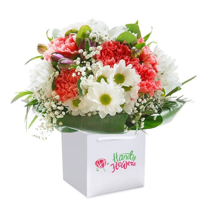 Finely presented in a modern, square white gift bag, this vibrant floral bouquet radiates freshness and warmth, inviting a sense of celebration that echoes throughout Longford's friendly community. At its heart, coral-pink and deep red carnations display their trademark ruffled petals, each layer capturing delicate highlights in the soft, diffused light, while cheerful white daisies with lime-green centers bring an uplifting contrast reminiscent of the meadows near Bath Road. Sprigs of feathery baby's breath meander through the bouquet, imparting an ethereal, cloud-like texture that softens the arrangement and creates depth. Blushing purple-tinged buds nestle subtly among the larger blooms, suggesting unfolding beauty and hinting at the promise of more to come. Surrounding it all, a generous array of deep green, broad leaves forms a natural, glossy collar, designed to showcase and cradle each blossom with equal care. The bouquet is carefully shaped into a full, rounded dome, conveying abundance and attention to detail, perfectly suited for everything from welcoming a new neighbour to marking an anniversary or birthday. The pristine white bag, finished with thick ribbon handles for easy delivery around Longford, highlights the arrangement's presentation-ready quality and ease of gifting. The entire composition glows against a pure white background, directing all focus to the exuberant colours and intricate textures. Crafted with local expertise, this bouquet reflects both the artistry and hospitality that define Longford's most cherished moments.