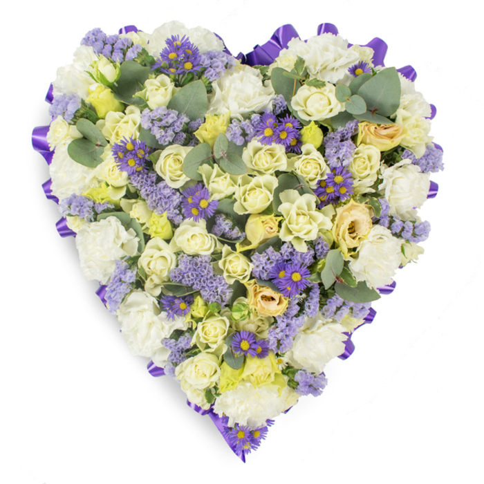 A meticulously constructed heart-shaped floral tribute rests against a bright white studio backdrop, its surface densely packed with soft, sympathetic hues that read as both tender and composed. The main body is woven from cream and pale yellow roses - varying from tight buds to open blooms with a faint green cast on some petals - lending the arrangement a layered, plush texture that feels quietly luxurious. Nestled among these are abundant white carnations, their ruffled petals creating cushiony swathes of gentle cream that support and amplify the roses' presence. Intermittent sprays of lavender statice bring a delicate, dusty-lilac shimmer across the design, while small, purple daisy-like asters with strong yellow centres create focused accents of deeper colour that catch the eye without breaking the arrangement's calm. Blue-green eucalyptus leaves, rounded and subtly waxy, are placed throughout to introduce a cool, architectural contrast and to suggest a fresh, balsamic fragrance. A continuous deep purple satin ribbon traces the heart's outer edge in neat, folded loops, framing the florals and giving the tribute a composed, finished silhouette suitable for memorial display. The lighting reveals petal detail and soft shadows, emphasizing the dense craftsmanship and the arrangement's purpose: a symbol of remembrance and enduring affection. This piece is particularly fitting for funeral services, chapel tributes and graveside placement in Longford town - a comforting expression of sympathy for families gathering at local venues or at the Market Square for a private farewell.