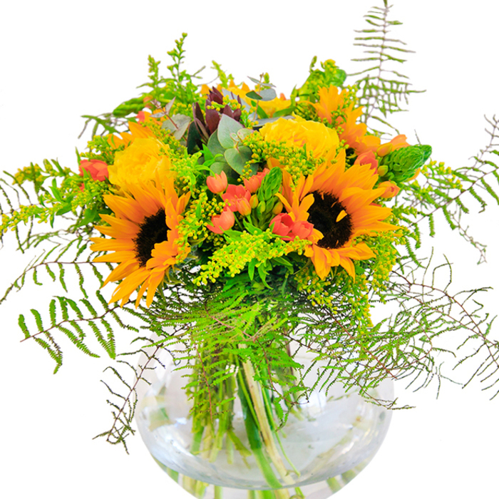 The first impression from this lush bouquet is pure joy-the vivid yellows and warm oranges of sunflowers and garden roses brightening the room like sunshine streaming through the windows of a Longford cottage. Arranged with a keen eye for texture and movement, the large, sun-drenched sunflower heads unfurl alongside clusters of classic garden roses, delicately packed with layers upon layers of petals. Touched with vibrant red Hypericum berries, reminiscent of the hedgerow fruits found in gardens near Connolly Barracks, the arrangement comes alive with contrasting highlights. The deep green foliage is a lush tapestry, with fine wisps of feathery fern and gentle hints of solidago, recalling the wildflowers that dance in the breeze along the Black River's edge. Slender flower spikes pierce the bouquet, adding vertical interest among brushing leaves of fresh eucalyptus that shimmer pale blue-green in the sunlight. The clear, rounded showcase vase reveals every detail: tangled stems immersed in sparkling water beneath a crown of blooms-capturing the artistry of local florists in Longford. Up close, every element invites touch and admiration, from the suede-like sunflower faces to the crisp edges of newly cut greens. Subtle fragrance drifts upward-a gentle mix of fresh grass, blossoming petals, and sun-warmed fields. Perfect as a bright centrepiece for a family table or as a gesture of celebration, the bouquet embodies the generous spirit of Longford, offering a heartfelt moment of connection and happiness.