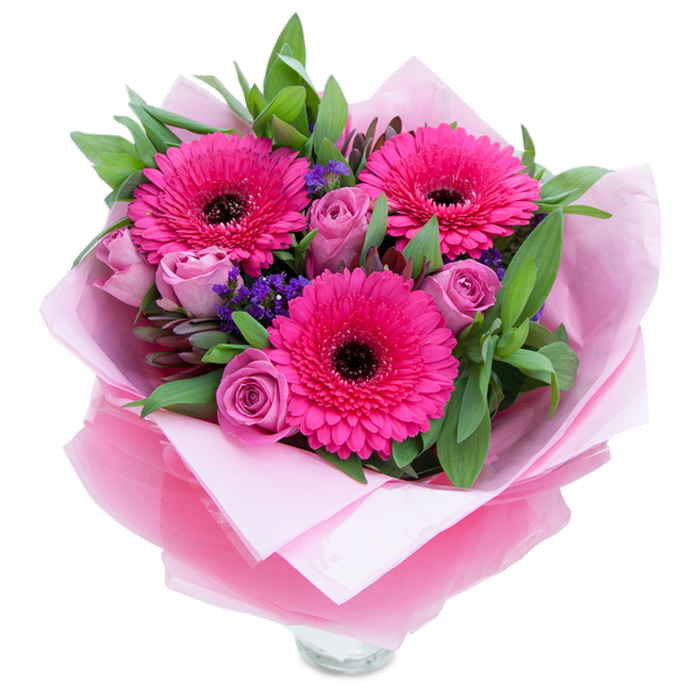 There is a playful grace to this bouquet, beginning with three confident fuchsia Gerbera daisies whose saturated petals fan outward like small suns, each centre a bold, dark counterpoint that anchors the composition. Nearby, roses in a muted lavender-pink or dusty-rose unfurl with refined curves, their tighter buds hinting at slow-opening beauty. Sprigs of purple statice or limonium are tucked through the stems, lending a papery texture and cool, jewel-toned punctuation against the pinks. The green foliage-broad salal leaves paired with the rounded, silvery sheen of eucalyptus-wraps the flowers in a garden-fresh embrace and gives the bouquet a pleasing, natural weight. It's presented with an inner layer of soft pale pink tissue, then finished with a sheer, crinkled fuchsia-pink cellophane that creates a subtle sparkle and keeps the colour story cohesive; the base shows the rim of a clear glass vase, signalling immediate display. In a quiet kitchen flooded with afternoon light the arrangement reads as intimate and cheerful at once: the roses breathe a gentle perfume, the gerberas offer a faint green note, and the statice adds a whisper of rustic charm. Thoughtfully crafted for occasions from birthdays to quiet celebrations, this bouquet carries the hospitality of local florists who know Longford's streets and homes-meant to be delivered, unwrapped, and enjoyed by someone special.