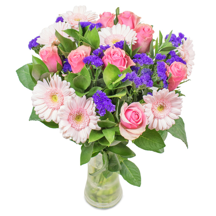 A graceful floral arrangement crafted in Longford, this bouquet boasts a captivating medley of soft pink roses-each bloom displaying its iconic spiral formation, some still shyly closed, others just beginning to expand. Interwoven are the distinct, round faces of gerbera daisies, their subtle shades of blush and pink glowing under natural light, the petal tips feathered in the gentlest cream. Vivid purple statice, resembling sprays of wildflowers from the fields surrounding Longford Village Green, offers a striking counterpoint, its subtle texture drawing the eye and heightening the visual interest of the bouquet. Lush, waxy green foliage encircles and frames the central blooms, evoking the freshness and invigorating scent of local woodlands after spring rain. The sleek glass vase is both contemporary and timeless, echoing the clarity and purity of freshly gathered water. Visible stems and bubbling water contribute an authentic touch, hinting at the bouquet's recent arrival from the florist's workbench to your home. Suitable for a variety of moments-Mother's Day breakfasts, birthday gifts, or just because-this harmonious blend of roses, gerberas, and statice brings a sense of celebration and heartfelt connection to any Longford residence or office, filling the air with soft fragrance and tender sentiment.